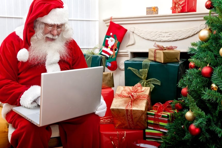 Santa Claus sitting next to a Christmas tree with a laptop