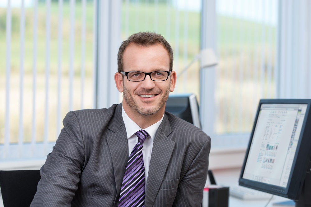 Friendly businessman wearing glasses sitting at his desk in the office looking at the camera with a smile.jpeg